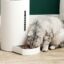 Gray cat eating from a modern smart pet feeder with automatic food dispenser on a wooden floor.